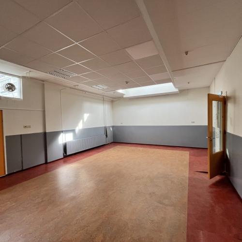 Grote, lege zaal met lichtgrijze muren, een oranje deur links en een houten deur rechts.
