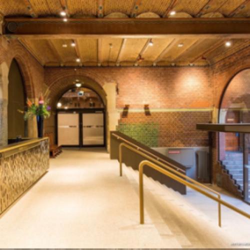 Warme hal met baksteenarchitectuur en houten balken. Links staat een balie en rechts is een verlaagde gang afgebakend door een boog en leuning.