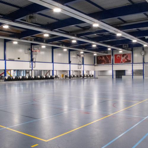 Groot, leeg sportgymzaal met een geel en blauw gemarkeerde vloer en ballenrekken aan de wanden.