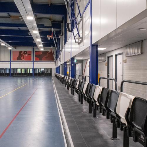 Lege gymzaal met een sportvloer en een lange rij lege stoelen aan de rechterkant.