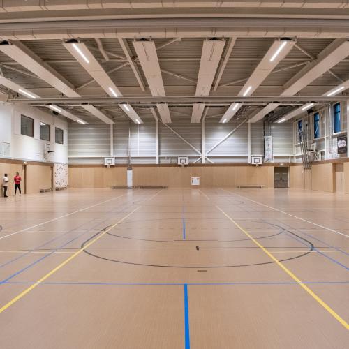 Binnenplan van een lege sporthal met een houten vloer en sportmarkeringen; lichte, industriële sfeer.
